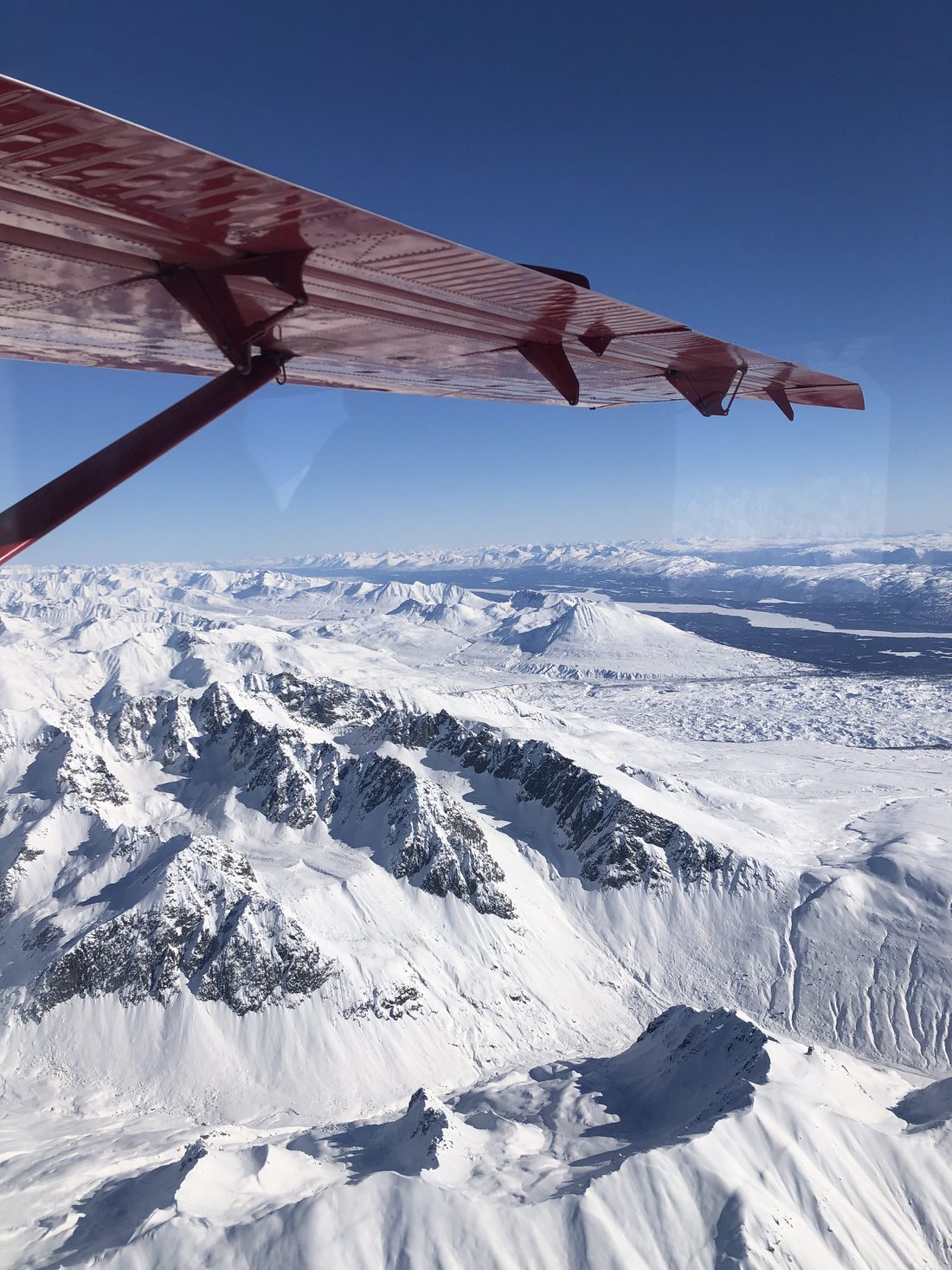 View past a red wing strut over snow-covered mountains and a glacier