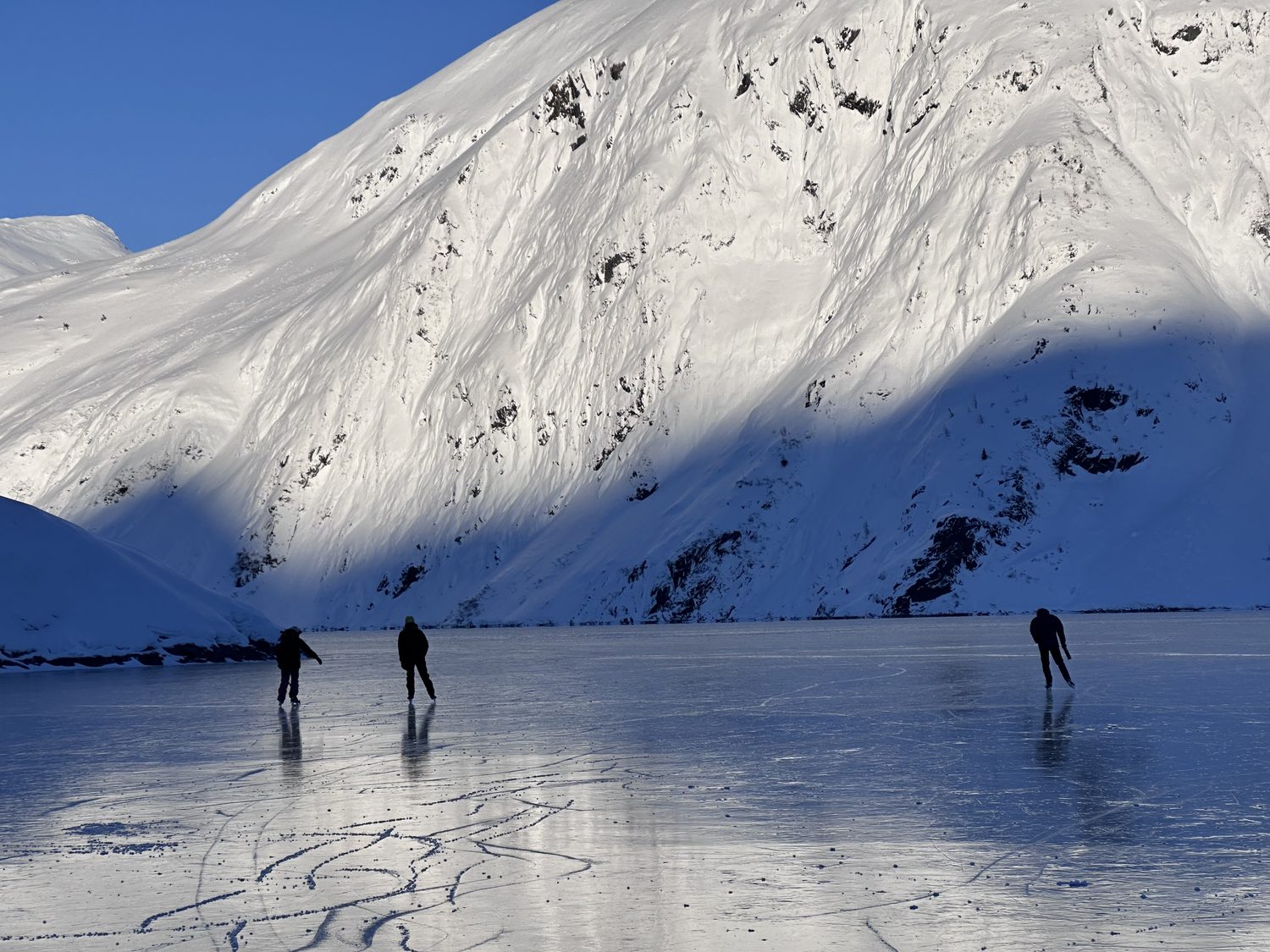 Three skaters on a frozen lake beneath a steep snow-covered slope